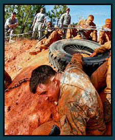 Service members competing in a team obstacle course moving a tire through the mud.
